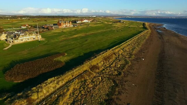 Aerial Moving Forward Across The Lush Greens Of The Royal Troon Golf Club, With Blue Skies, Historic Buildings, And The Rugged Coastline Stretching To The Horizon