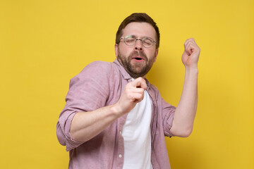 Fototapeta premium Caucasian man in a good mood, he dances making movements with hands and sings along with eyes closed. Yellow background.