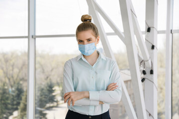 Young beautiful businesswoman in a protective medical mask stands near the window.