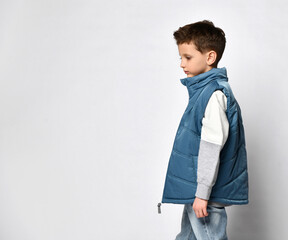 Side view of a teenage boy in a blue puffed sleeveless jacket, standing half a turn towards us, smiling, against a light studio background