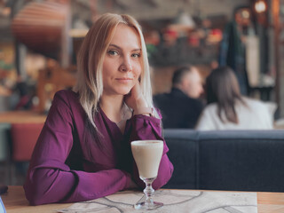 A blonde girl in a lilac sweater sits in a cozy restaurant on a sofa and she is relaxed and smiling and have a coffee drink on her table.