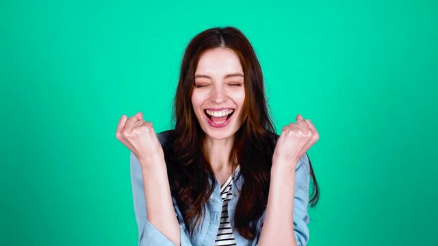 Happy, joyful, enthusiastic young girl with long dark hair, in trendy modern casual clothes, standing against a green background, clapping her hands, rejoicing in victory, good news.