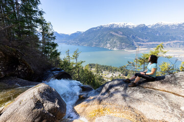 Adventurous Adult Woman is sitting on top of a Beautiful Waterfall, Shannon Falls, and watching the view. Taken near Squamish, North of Vancouver, British Columbia, Canada.