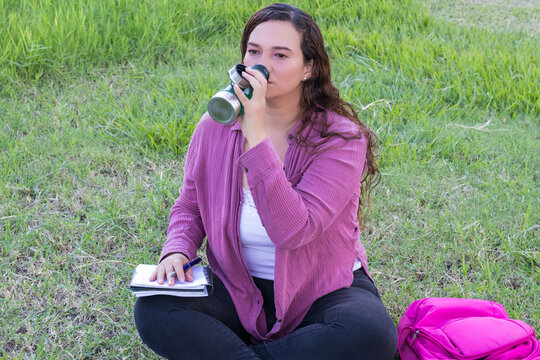 Hermosa joven estudiante colombiana sentada en el césped bebiendo agua con un cuaderno en su pierna