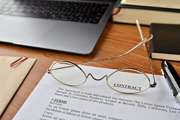 There is a dummy paper of Contract on the desk with a laptop, a pen and glasses.