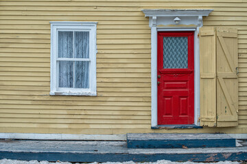 Fototapeta premium The exterior wall of a yellow wood clapboard heritage house. The door of the building is bright red with a small glass window. There's a lace curtain in the double hung window and an iron patio bench.