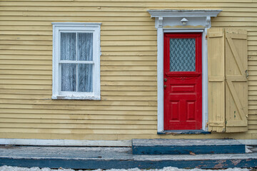 The exterior wall of a yellow wood clapboard heritage house. The door of the building is bright red with a small glass window. There's a lace curtain in the double hung window and an iron patio bench.