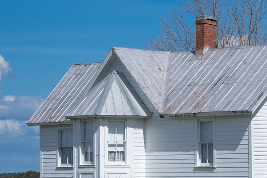 A white vintage building with white trim and dark roof shingles. The top of the building has a chimney and multiple old double hung windows. The background has blue skies and trees.