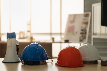 Closeup of blue, orange and white hard safety helmet hat of workman as engineer and ทegaphone on wooden table with building blueprint as background.