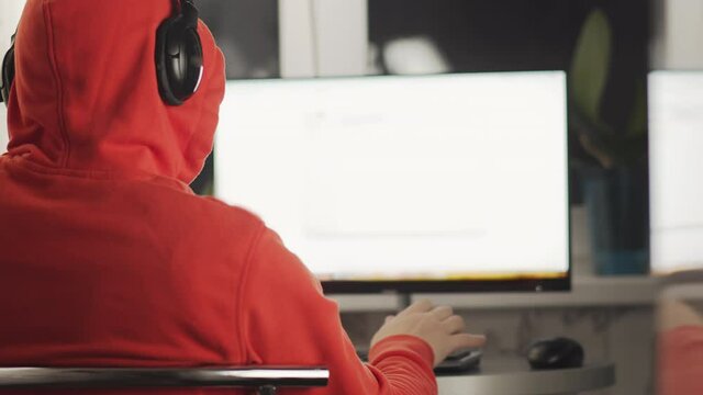 A programmer in a hood works in a home office in the evening sitting at a monitor screen, copyspace