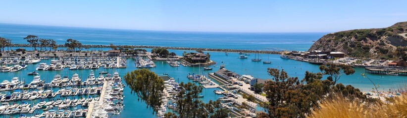 a stunning panoramic shot of the boats and yacht docked and sailing Dana Point Harbor with deep blue ocean water, lush green plants and trees on the hillside and blue sky in Dana Point California - obrazy, fototapety, plakaty