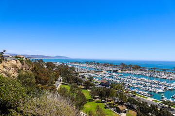 a stunning shot of the boats and yacht docked and sailing Dana Point Harbor with deep blue ocean water, lush green plants and trees on the hillside and blue sky in Dana Point California	 - obrazy, fototapety, plakaty