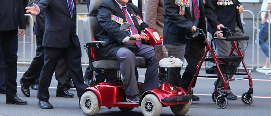 A group of older men wearing military medals and berets marching. one man is using a walker another is using a mobility scooter. People at the Anzac Day March, Sydney City.