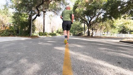 2020 - An old man jogs away from the camera in slow motion in the middle of a tree-lined street in Ojai, California.