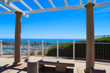 Fototapeta na wymiar a white wooden awning with stone pillars near a view of stunning deep blue ocean water with benches underneath and blue sky and lush green trees at Dana Point Bluff Top Trail in Dana Point CA