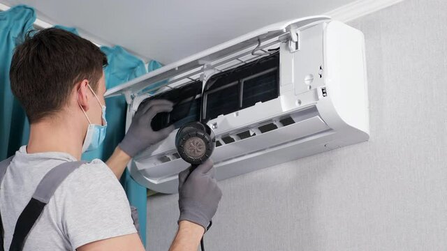 Concentrated repairman in gray t-shirt and face mask cleans ceiling air conditioner unit with dryer near azure curtains in room closeup