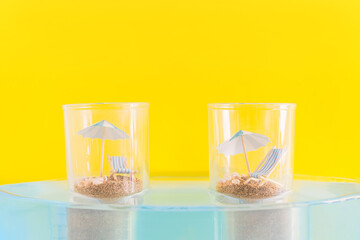 Summer vacation concept during pandemic. Social distance of people on the beach. Two glasses with sand, chaise longue and beach umbrella standing in the water