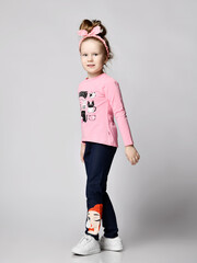 Blonde kid girl in comfortable summer clothing blue jeans with girlish print, pink long sleeve t-shirt and sneakers is walking passing by camera over light background