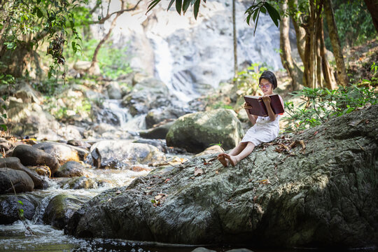 Asian little girl with glasses sitting on stone reading a books near forest waterfall
