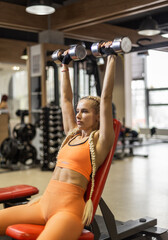 Fototapeta na wymiar Fitness woman in sportswear trains shoulder muscles, doing dumbbell press while sitting on bench in gym