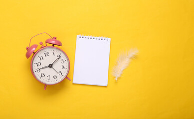 Feather and notebook with alarm clock on yellow background. Minimalism. Top view. Flat lay