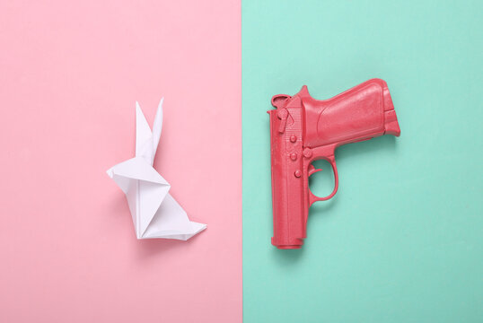 Pink pistol gun and origami rabbit on blue pink background. Creative concept. Minimalism Flat lay. Minimal layout.Top view