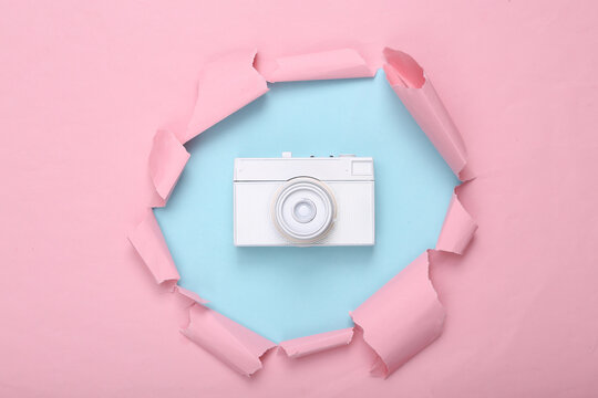 Creative layout. White retro camera through torn hole of pink blue paper. Pastel color trend. Fresh idea. Minimalism. Concept art. Flat lay. Top view.