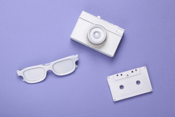 Minimalistic layout of white audio cassette, glasses, camera on purple background. Top view. Flat...