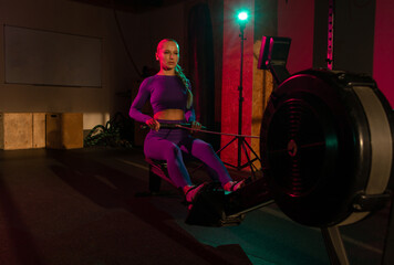 Fototapeta na wymiar Woman doing cardio workout on rowing machine in pink green neon light at gym. Sportive girl works out in fitness club. Healthy lifestyle