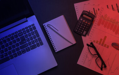Laptop, classic eyeglasses, calculator, notebook with graphs and charts in neon light. Economic...