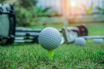 Closeup golf ball on tee ready to be shot. Golf ball on tee in the evening golf course with sunshine. Blurred set of golf clubs over green field background.