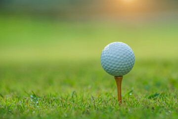 Closeup golf ball on tee ready to be shot. Golf ball on tee in the evening golf course with sunshine. Blurred set of golf clubs over green field background.