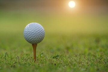 Closeup golf ball on tee ready to be shot. Golf ball on tee in the evening golf course with sunshine. Blurred set of golf clubs over green field background.