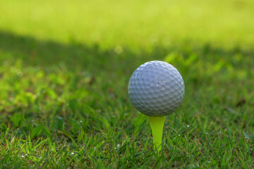 Closeup golf ball on tee ready to be shot. Golf ball on tee in the evening golf course with sunshine. Blurred set of golf clubs over green field background.