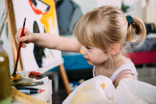 A little cute girl paints a big picture with acrylic at home on an easel. 