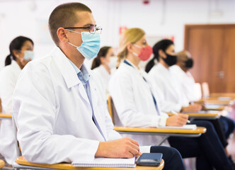Fototapeta premium Focused man in white coat and protective mask sitting in conference room during medical seminar. Necessary precautions in coronavirus pandemic