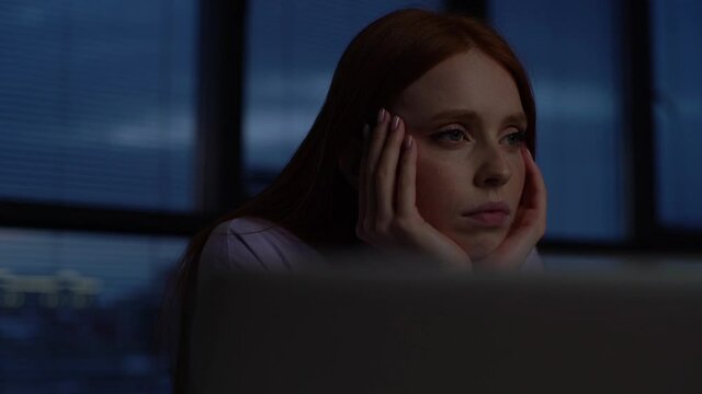 Close-up face of tired sad woman sitting at desk with laptop in dark office room near window at night. Tracking shot of depressed upset female thinking about mistakes in work and looking away at home.