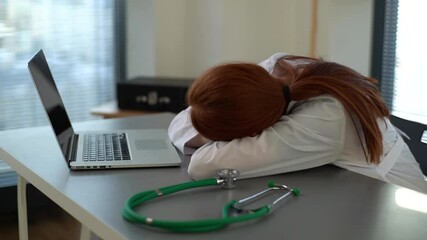 Close-up of tired overworked redhead female doctor in white coat sleeping at desk with laptop and stethoscope on workplace near window in office room of medic clinic. Tracking shot in slow motion. - Powered by Adobe