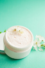 Fototapeta na wymiar Jar of white skincare cream with Fresh spring cherry blossom flowers. Cosmetic bottles. Botanical spa treatment delicate skin. turquoise background