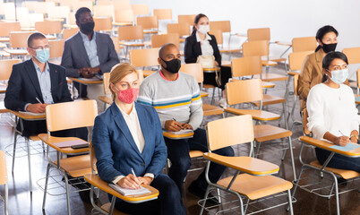 Fototapeta na wymiar Multiethnic group of people wearing protective masks sitting in conference room keeping distance during business training. Precautions during mass events in coronavirus pandemic