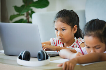 Two asian child girl students study online with teacher by video call together. Siblings are homeschooling with computer laptop during quarantine due to Covid 19 pandemic.