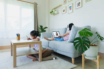 Asian kid sitting on sofa and having video conference chat with teacher and class group with sister doing worksheet stay beside. Children study from home during quarantine due to COVID-19 outbreak.