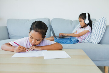 Asian child girl doing worksheet with her sister sitting on sofa and having video conference chat with teacher and class group. Children are homeschooling during quarantine due to COVID-19 outbreak.