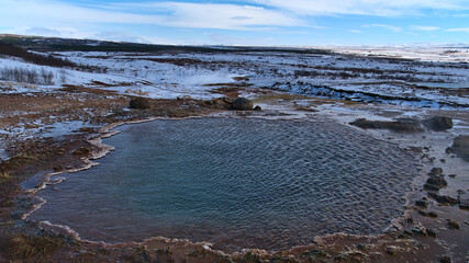 Hot spring with steaming pool of water in Geysir in geothermal area Haukadalur, part of Golden Circle, Iceland in winter season on sunny day with snow-covered landscape.