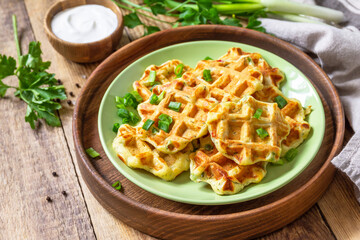 Not sweet waffles with potatoes, peas, bell peppers, cheese are served with sour cream and herbs on a plate on a rustic table.