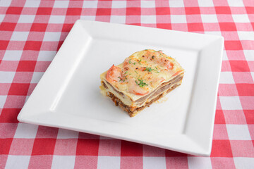 One piece of Italian lasagna on a square white plate over red plaid table cloth.