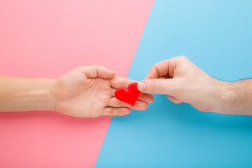 Young adult man hand giving red heart shape to woman palm on light pink blue table background. Pastel color. Concept of fall in love. Closeup. Lovely emotional, sentimental life moment. Top view.