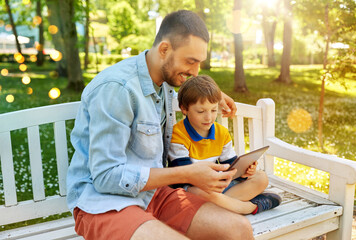 family, fatherhood and technology concept - happy father and little son with tablet pc computer sitting on bench at summer park