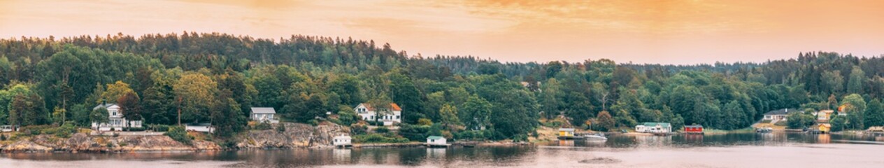 Fototapeta na wymiar Sweden. Many Beautiful Swedish Wooden Log Cabins Houses On Rocky Island Coast In Summer Evening. Lake Or River Landscape. Panorama, Panoramic View.