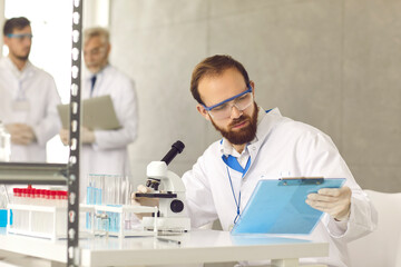 Naklejka na ściany i meble Portrait of man scientist in eyeglasses and lab coat sitting at desk looking at clipboard with test result analyzing sample using microscope. Research group working at laboratory on background